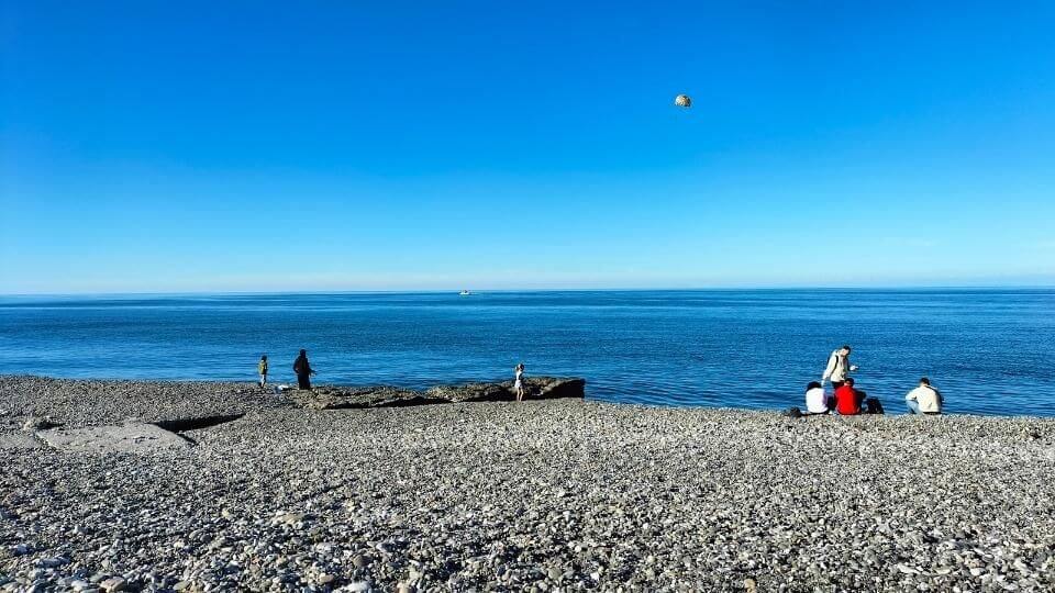 People sitting on the pebble beach with parasailers in the sky overhead on a stunning blue-sky day in Batumi, Georgia.
