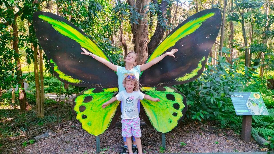 Ayla and Romy stretch their arms out in front of butterfly wings at Indigiscapes, one of our recommended attractions for families in Brisbane.