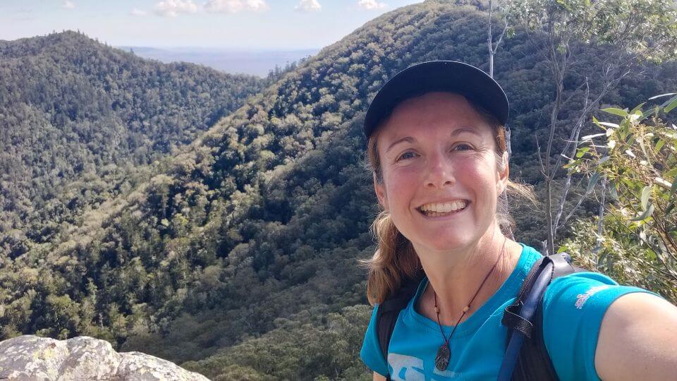 Elly takes a selfie at the top of Mount Woowoonga, with a nice view of lush green hills behind.