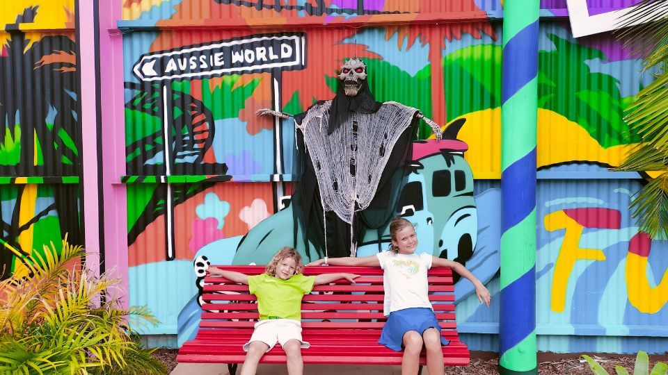 Ayla and Romy sit on a bench in front of a colorful wall at Aussie World, complete with Halloween decor.