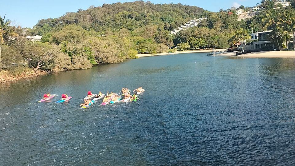 Noosa surf club members train on their boards on the Noosa River, surrounded by stunning greenery.