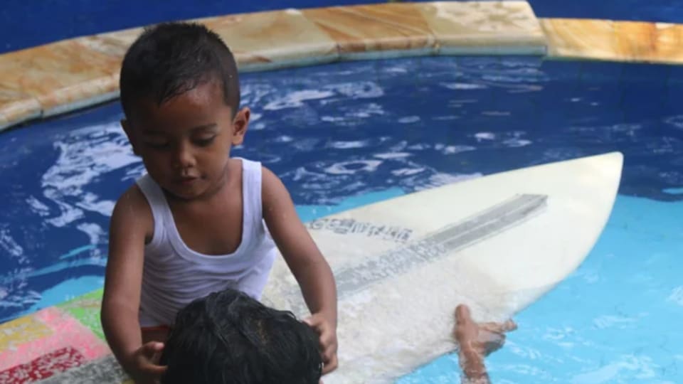 A kid sits on a surf board in a swimming pool while an adult holds the board at Pelan Pelan Surf & Yoga Retreat in Bali.