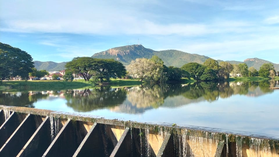 A picturesque shot of the Aplins Weir on the Ross River in Townsville with Castle Hill in the background, and trees reflecting onto the water.
