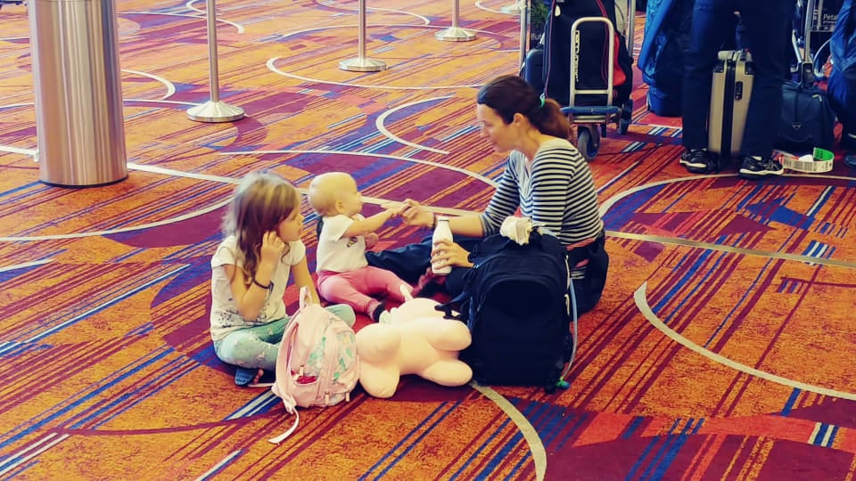 Ayla, Romy, and Elly sit on an airport floor with their carry-on luggage.