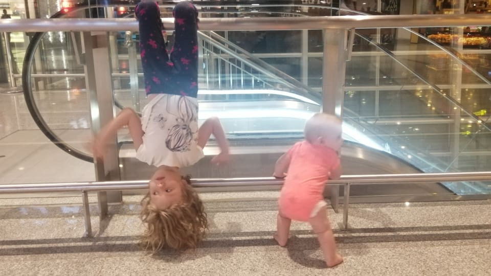 Ayla hangs upside down on a railing beside the top of an escalator, while baby Romy holds onto a lower railing.