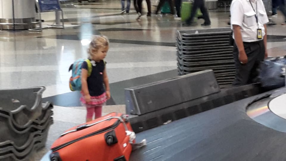 Ayla waits by the luggage conveyor belt at an airport, ready to collect bags.