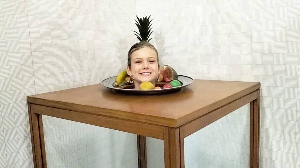 Ayla's head pops out of a fruit bowl on a table at an illusion exhibit in the science room of the Queensland Museum Tropics.