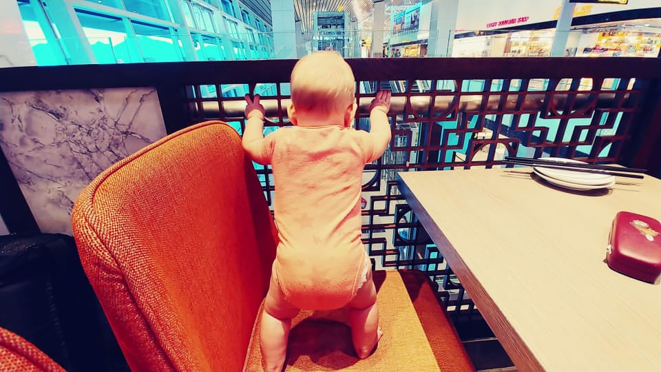 Baby Romy stands on a chair and holds onto a railing at an airport.
