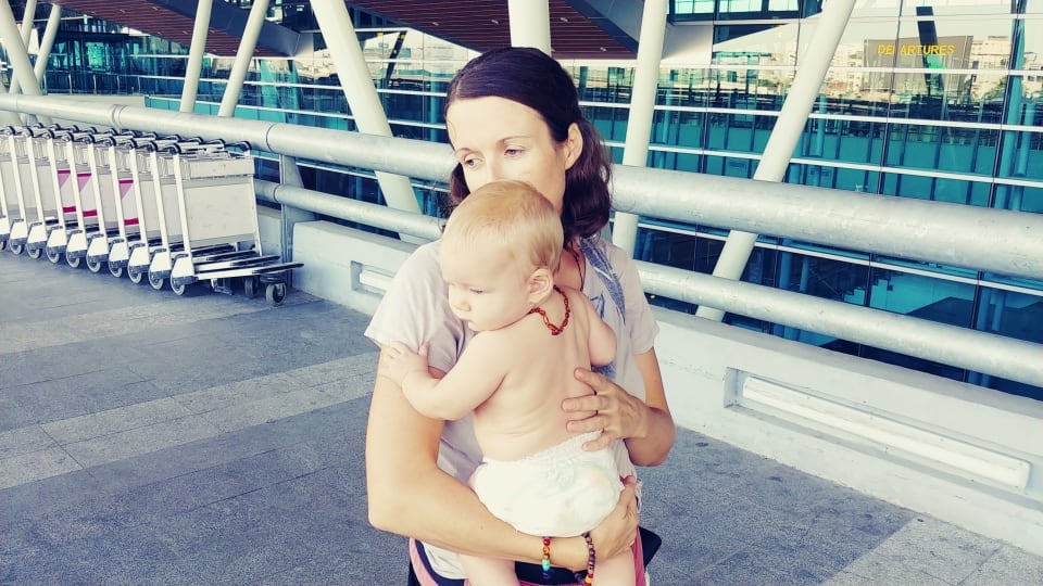 Elly holds baby Romy outside an airport near the luggage trolleys.