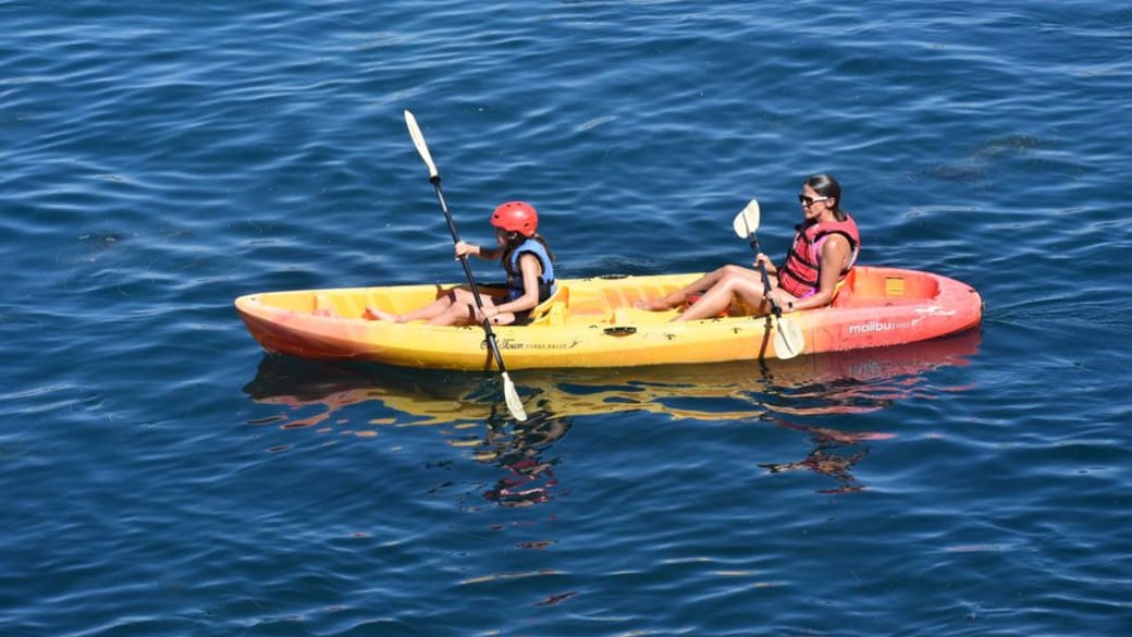 A mother and daughter paddle a kayak across a calm lake under clear skies.