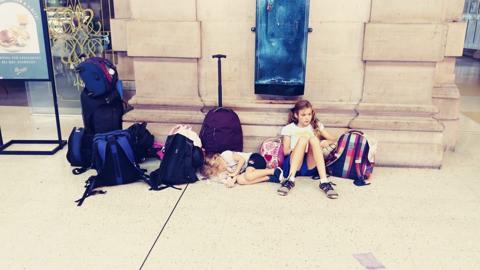 Ayla and Romy sit against a wall in a train station in Australia with all the family's luggage.