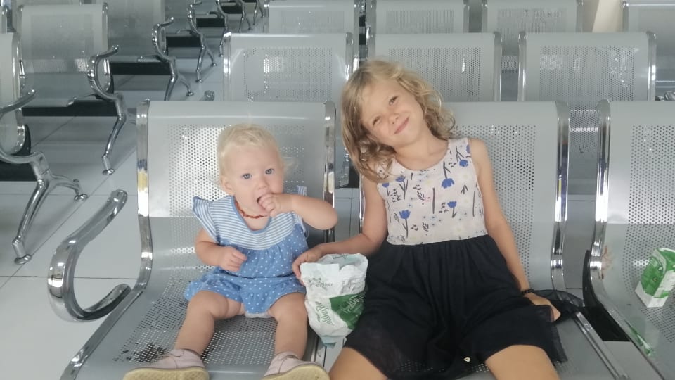Ayla and Romy sit on seats in a waiting area and eat some snacks on a family travel day.