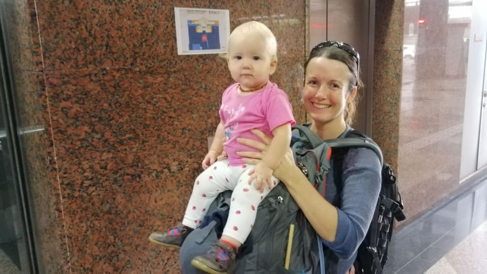 Elly carries a backpack on her back and front, and baby Romy on the top of the front pack in an airport on a family travel day.