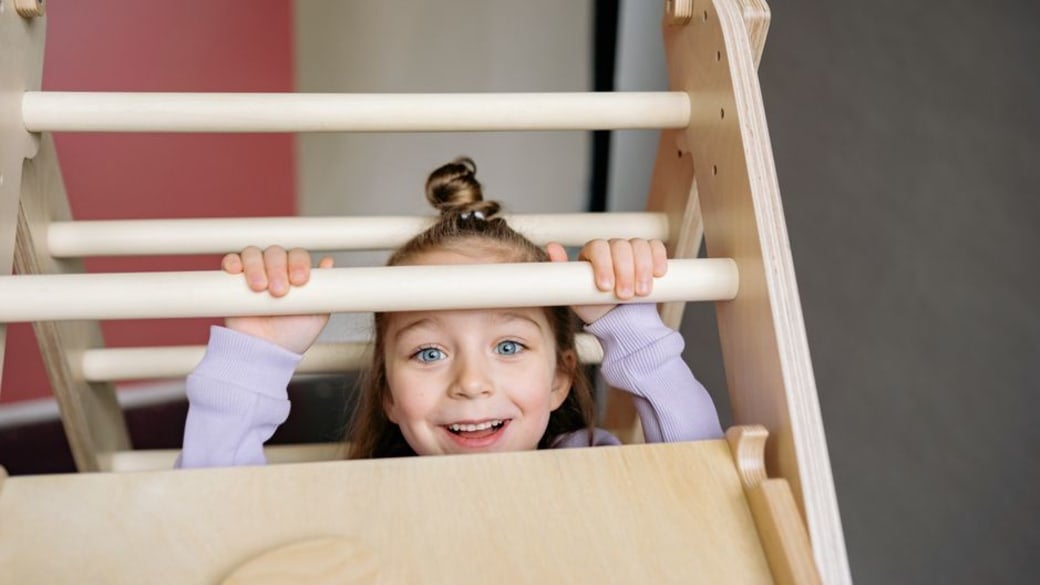 Cheerful little girl climbing on an indoor wooden play structure, beaming with happiness.