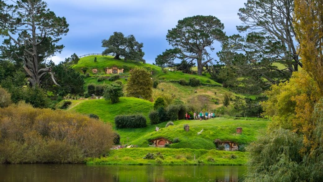 Lush green landscape of Hobbiton with iconic hobbit holes and serene lake reflecting the vibrant scenery.