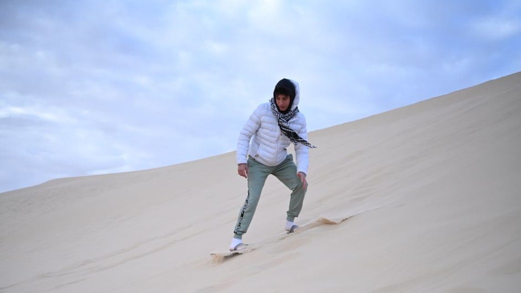 A man in a white jacket sandboarding on the vast desert sand dunes under a cloudy sky.