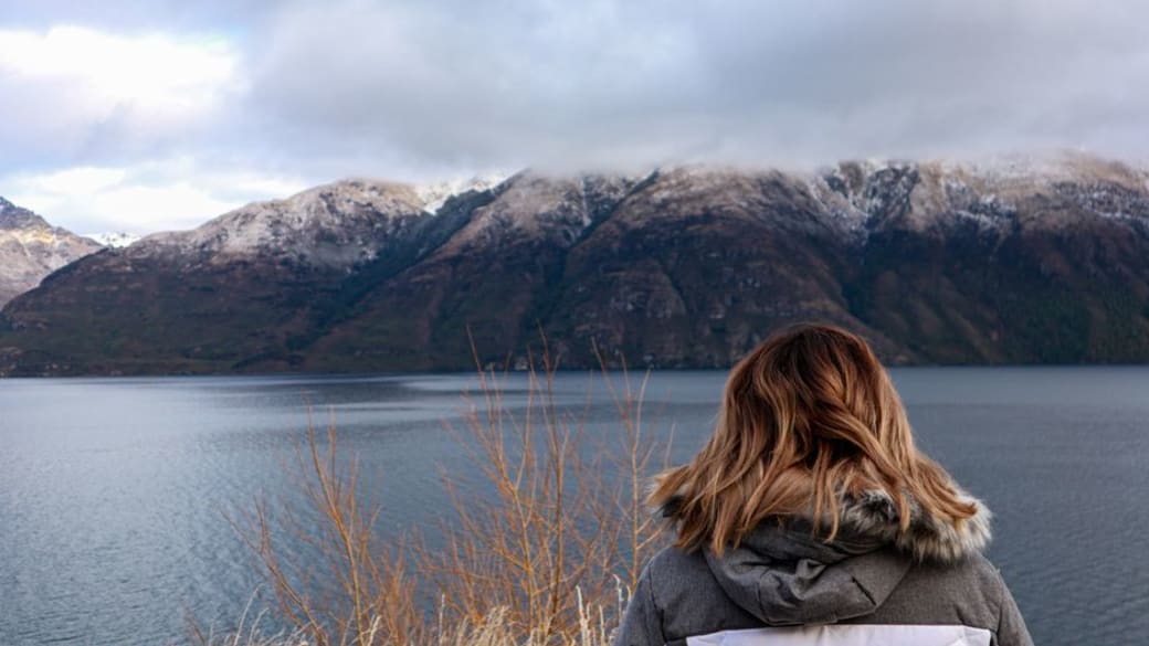 A woman with brown hair in a jacket gazes at Lake Wakatipu and snow-capped mountains in Queenstown, New Zealand.