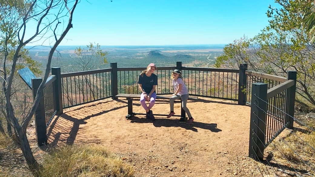 Ayla and Romy sit on a bench at a fenced lookout up Mount Zamia, with gorgeous views beyond.