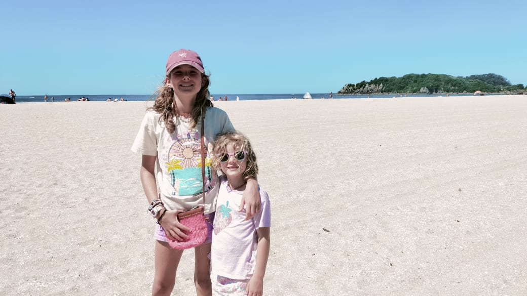 Ayla and Romy smile for a photo on Mount Maunganui beach near Tauranga, with Moturiki Island in the background.