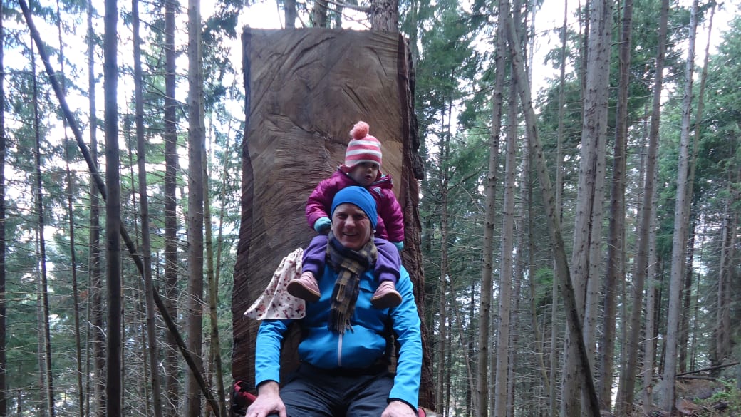 Colin sits on a big wooden chair in the forest in Queenstown with little Ayla on his shoulders.