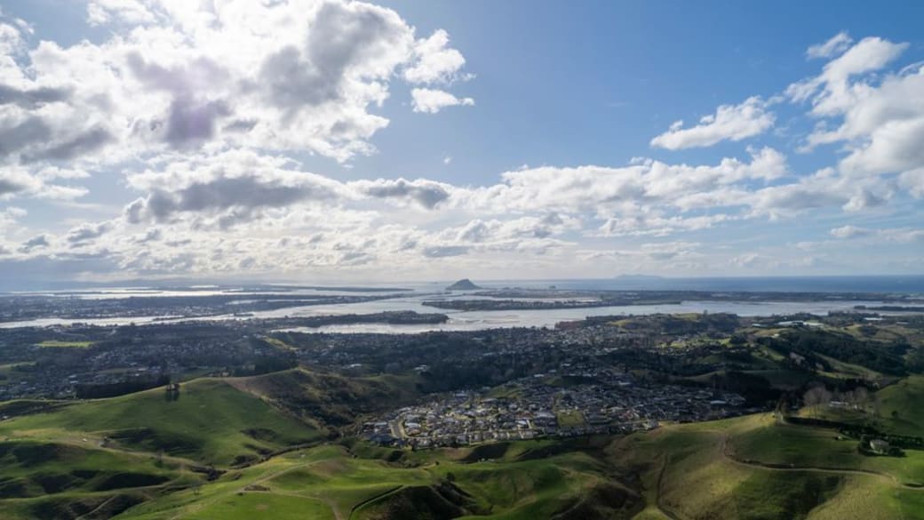 Stunning aerial view of Tauranga, New Zealand with hills, coastline, and clouds under a bright sky.