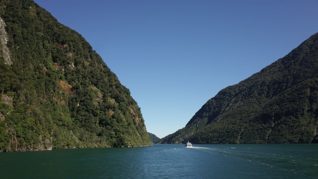 A boat navigates through the scenic waterway of Fiordland National Park, surrounded by lush green mountains.