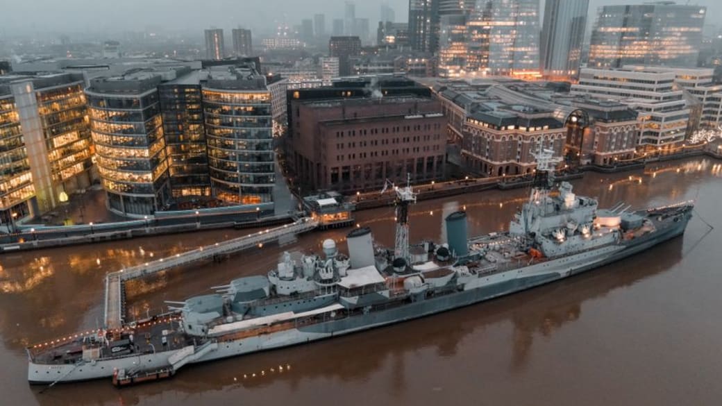 Aerial shot of the HMS Belfast moored on the Thames against London's illuminated skyline at dusk.