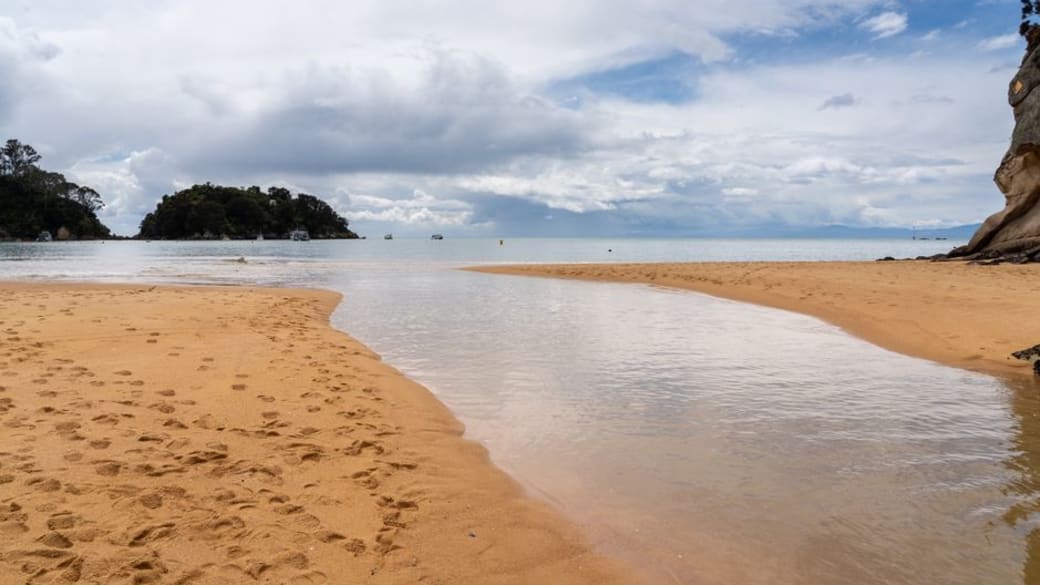 Stunning coastal view of a tranquil beach near Nelson NZ with golden sands, calm sea, and distant islands under a cloudy sky.