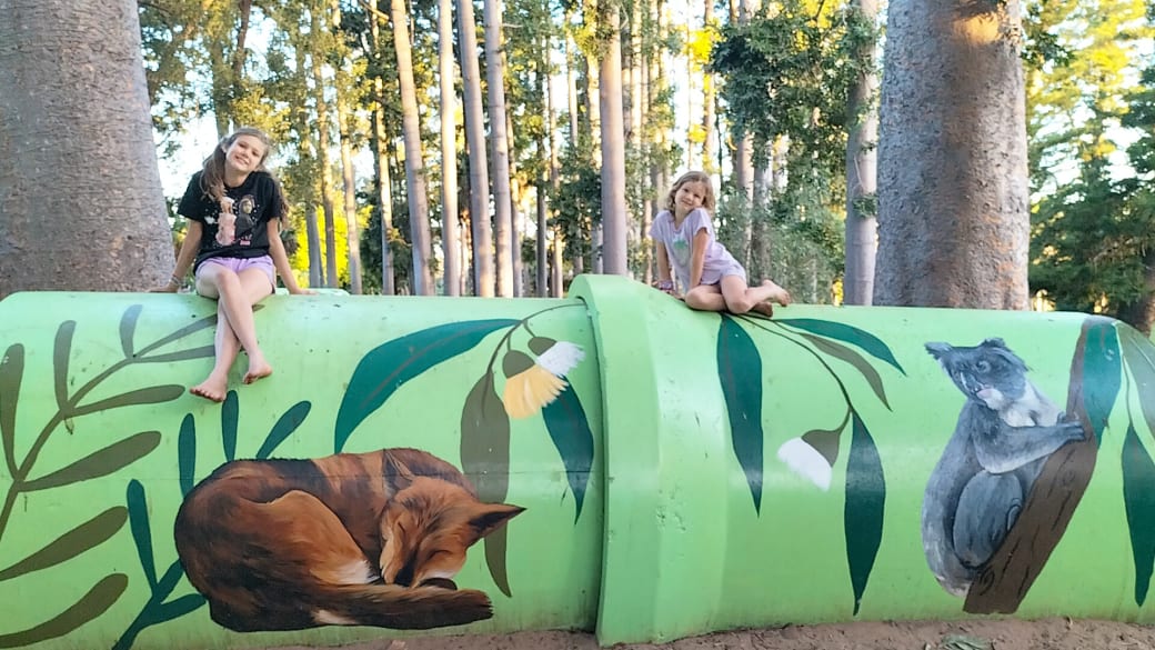 Ayla and Romy sit on top of colorful artwork in the Rockhampton Botanical Gardens with the sun shining through the trees behind.