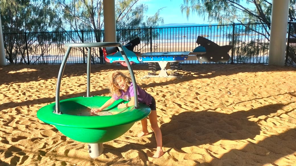 Romy in the sandy enclosed playground at Yeppoon Rotary Park, with Farnborough Beach in the background.
