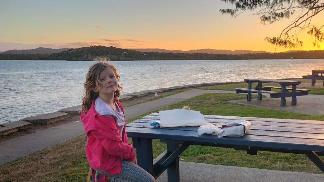 Romy sits at a picnic bench with fish and chips on the table and a beautiful sunset behind at Causeway Lake on the Capricorn Coast.